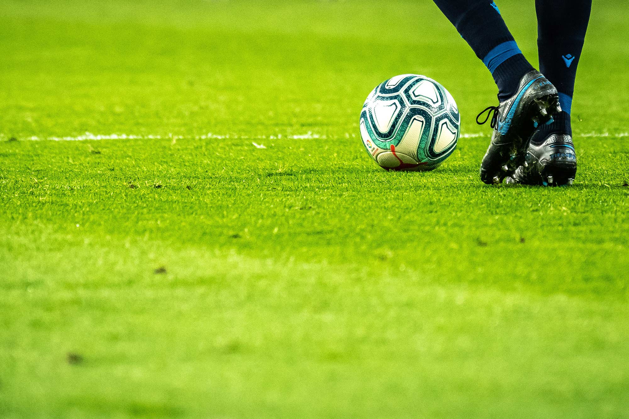 Soccer ball resting on a green soccer field as a player's feet wait to kick it