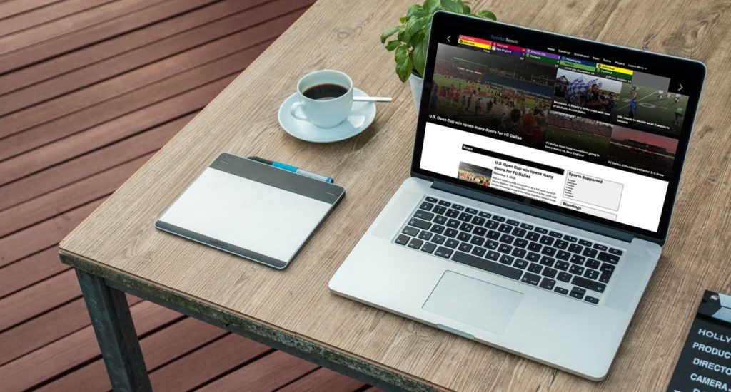 Apple MacBook Pro laptop with Sports Bench 2.0 on the screen sitting on a wooden table along with a notebook, cup of coffee and small plant